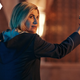 Jodie Whittaker's 13th Doctor has her back to the camera with her head turned and holds up a small, triangular beacon device.
